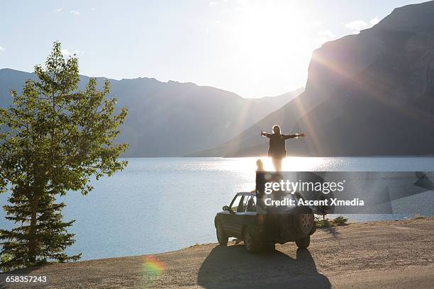 woman extends arms on car roof while man relaxes - distancia media fotografías e imágenes de stock