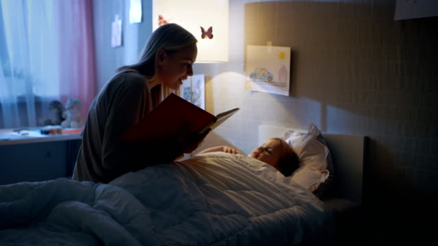 Young Loving Mother Reads Bedtime Stories to Her Little Beautiful Daughter who Goes to Sleep in Her Bed.
