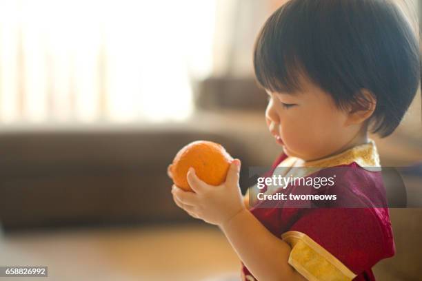 asian toddler in chinese traditional clothing cheongsam, holding tangerine. - tangerine 2015 film stock-fotos und bilder