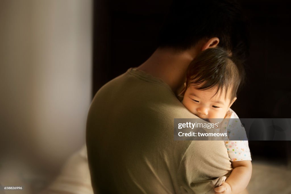 Asian toddler carried by father in moody bedroom background.