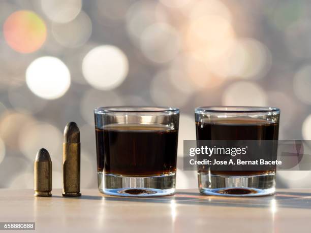 glass of crystal of chupito fill of an alcoholic drink and a bullet of pistol, , illuminated by the light of the sun - alcoholismo stock pictures, royalty-free photos & images