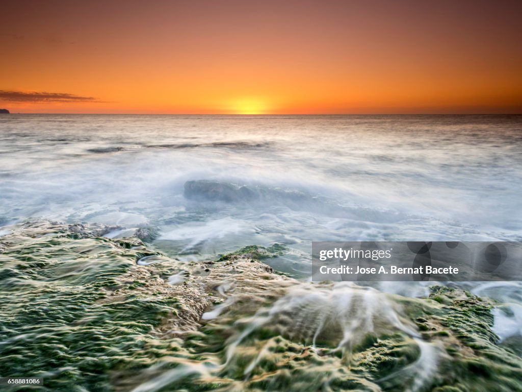 Exit of the Sun of orange color, on the surface of the sea, in a zone of coast with rocks and waves in movement