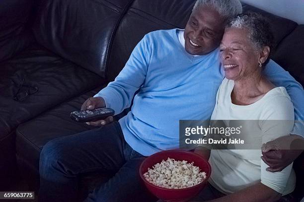 senior couple sitting on sofa at night, watching television, woman holding bowl of popcorn - watching tv couple night stock pictures, royalty-free photos & images