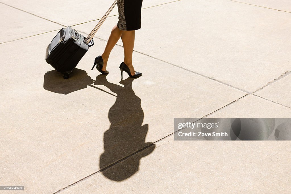 Businesswoman walking outdoors, wearing high heeled shoes, pulling wheeled suitcase, low section