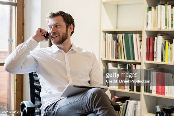 mid adult businessman using digital tablet and talking on mobile phone in an office, bavaria, germany - formal office stock pictures, royalty-free photos & images
