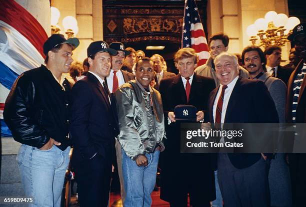 American businessman Donald Trump holding a New York Yankees baseball cap at an event in New York City, circa 1990. With him are boxer, Sugar Ray...