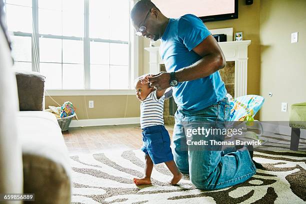 father helping infant son learn to walk in home - leanincollection father stock-fotos und bilder