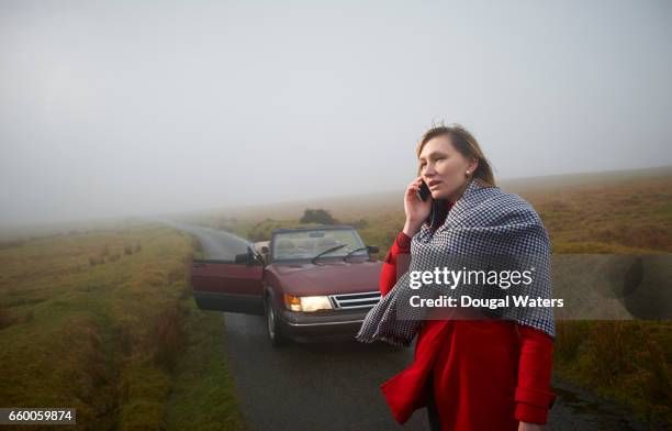 woman standing in road and making phone call with car in background. - car problems stock pictures, royalty-free photos & images