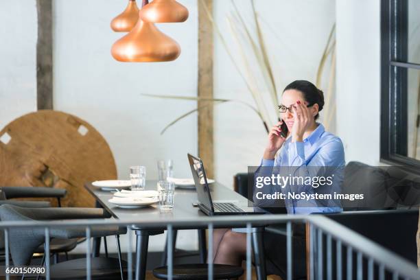 schöne brünette geschäftsfrau auf mittagspause, arbeiten am laptop im restaurant. sie trägt brille, im gespräch mit handy - brunette sitting at desk stock-fotos und bilder