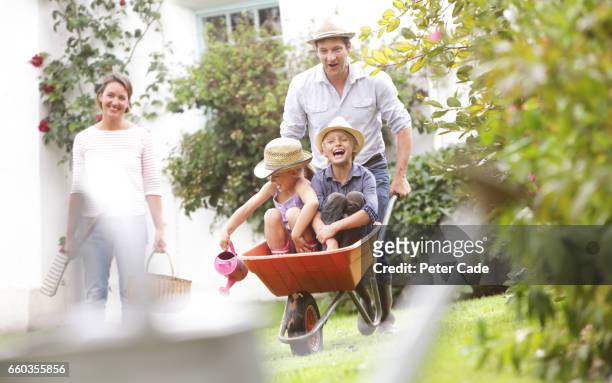 family playing in garden with children in wheelbarrow - family gardening stock-fotos und bilder