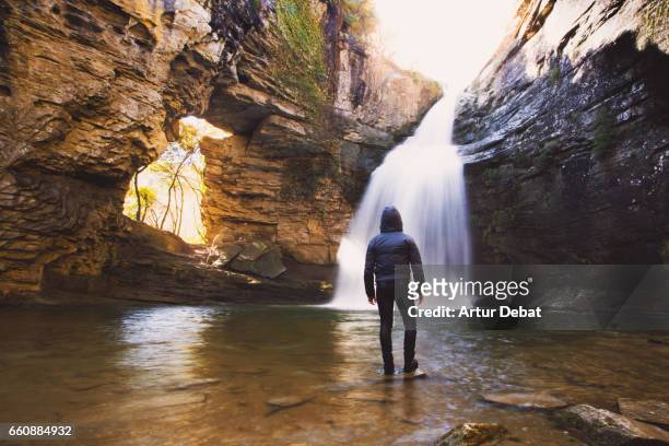 guy standing in the middle of the water contemplating the stunning and powerful waterfall falling from unique place with hole and natural arch in the mountain region of collsacabra with long exposure and amazing light. - uomo incappucciato foto e immagini stock