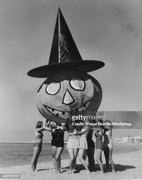 In advance of Halloween, a group of young women in swimsuit pose on the beach, a massive papier mache pumpkin in a witch's hat hoisted over their...