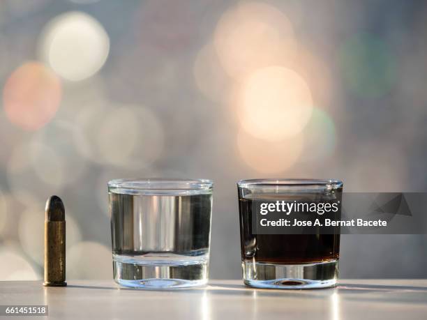 glass of crystal of chupito fill of an alcoholic drink and a bullet of pistol, , illuminated by the light of the sun - bebida stockfoto's en -beelden