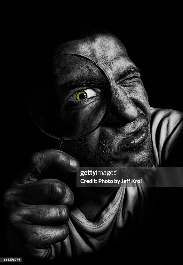 Man holding magnifying glass in front of his green colored eye. Investigating like a detective, looking into camera.