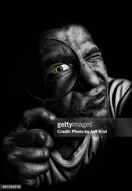 man holding magnifying glass in front of his green colored eye. investigating like a detective, looking into camera. - portretfoto imagens e fotografias de stock