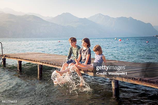 italy, brenzone, three children sitting side by side on jetty splashing with water - jetty stock pictures, royalty-free photos & images