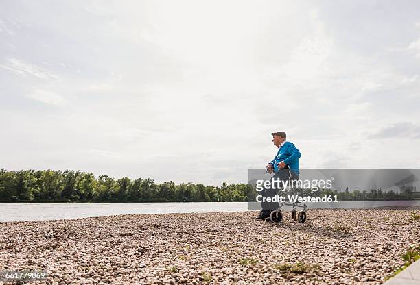 senior man sitting on wheeled walker at riverside - rollator stock-fotos und bilder