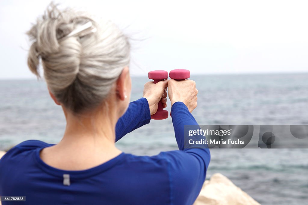 Mature woman beside sea, exercising with hand weights, rear view