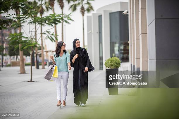 young middle eastern woman wearing traditional clothing walking along street with female friend, dubai, united arab emirates - middle eastern women foto e immagini stock
