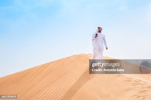 middle eastern man wearing traditional clothes using smartphone on desert dune, dubai, united arab emirates - arab man walking stock pictures, royalty-free photos & images