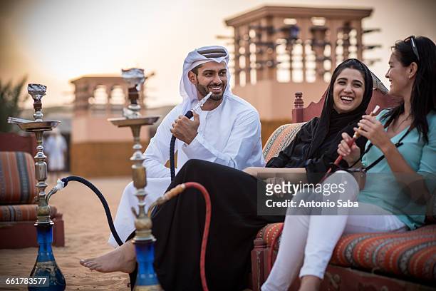 local couple wearing traditional clothes smoking shisha on sofa with female tourist, dubai, united arab emirates - dubai desert stock pictures, royalty-free photos & images