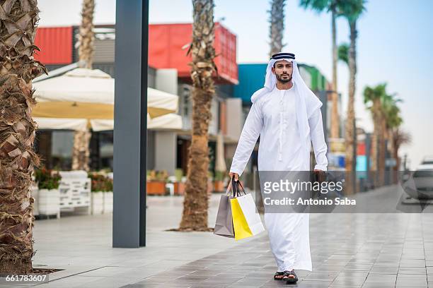 man wearing traditional middle eastern clothing walking along street carrying shopping bags, dubai, united arab emirates - arab shopping stockfoto's en -beelden