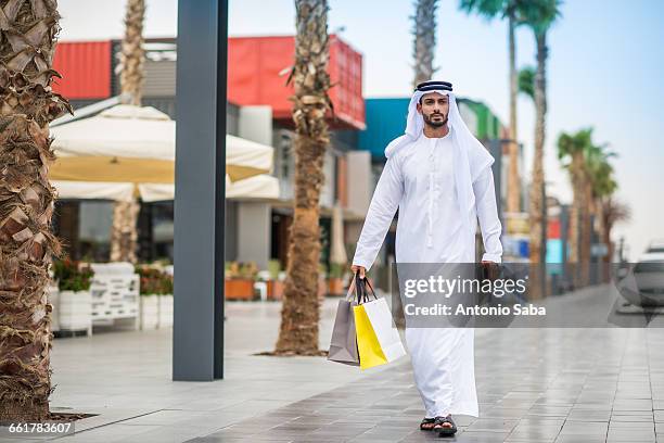 man wearing traditional middle eastern clothing walking along street carrying shopping bags, dubai, united arab emirates - arab man shopping stock pictures, royalty-free photos & images