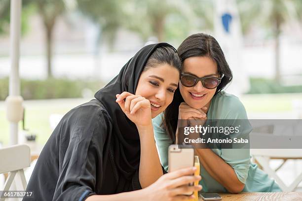 young middle eastern woman wearing traditional clothing reading smartphone text with female friend at cafe, dubai, united arab emirates - middle east friends stock pictures, royalty-free photos & images