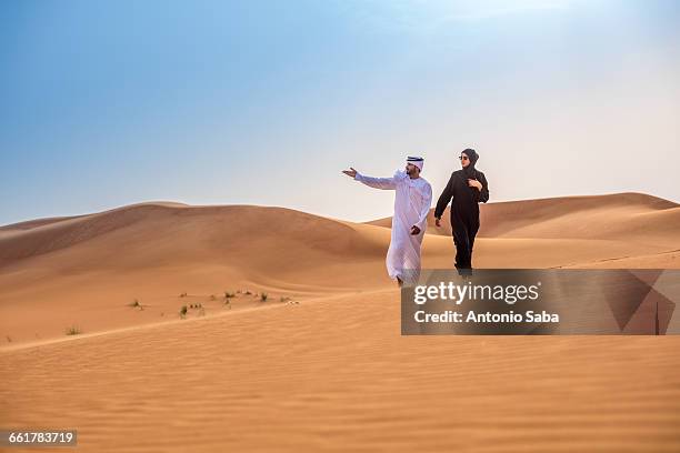 couple wearing traditional middle eastern clothes pointing from desert dune, dubai, united arab emirates - middle east clothing stock pictures, royalty-free photos & images