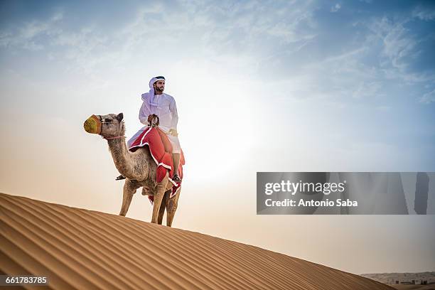 man wearing traditional middle eastern clothes riding camel in desert, dubai, united arab emirates - tradition stock pictures, royalty-free photos & images