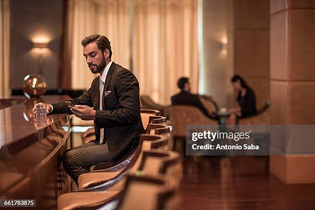 young businessman sitting at hotel bar reading smartphone texts - man in bar stockfoto's en -beelden