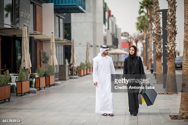 middle eastern shopping couple wearing traditional clothing carrying shopping bags, dubai, united arab emirates - arab woman walking stock-fotos und bilder