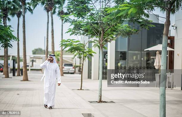 man wearing traditional middle eastern clothing walking along street talking on smartphone, dubai, united arab emirates - arab phone stock-fotos und bilder