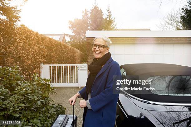 woman near car on driveway holding suitcase looking at camera smiling - garagentor stock-fotos und bilder