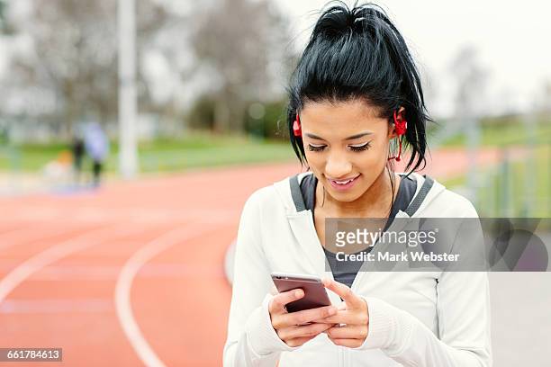 young woman beside running track, holding smartphone, wearing earphones, smiling - live at leeds 2016 stock pictures, royalty-free photos & images