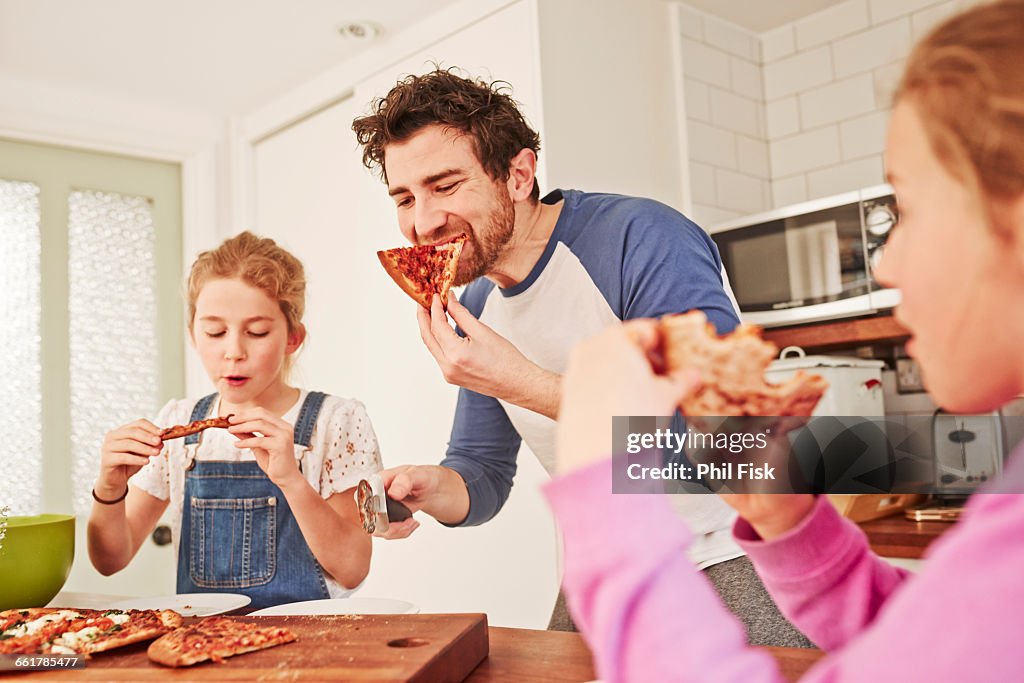 Mid adult man eating pizza with daughters at kitchen bench