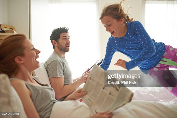 mid adult couple reading broadsheet whilst daughter leaps on bed - read and newspaper and bed stock pictures, royalty-free photos & images