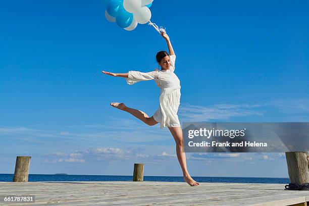 young woman dancing on wooden pier, holding bunch of balloons - white dress stock-fotos und bilder