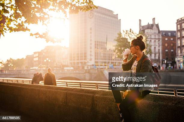 woman drinking coffee while sitting retailing wall of bridge in city - cosmopolitan drink stock pictures, royalty-free photos & images