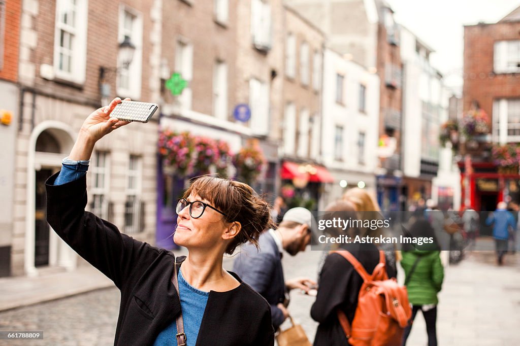 Smiling woman taking selfie with friends standing in background on city street