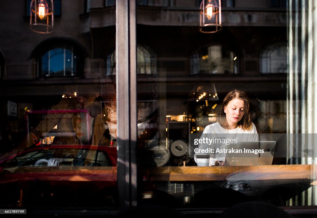 Woman using laptop in cafe seen through glass window