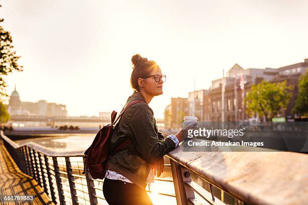 side view of woman holding disposable cup while standing on boardwalk by river - brille fluss stadt stock-fotos und bilder