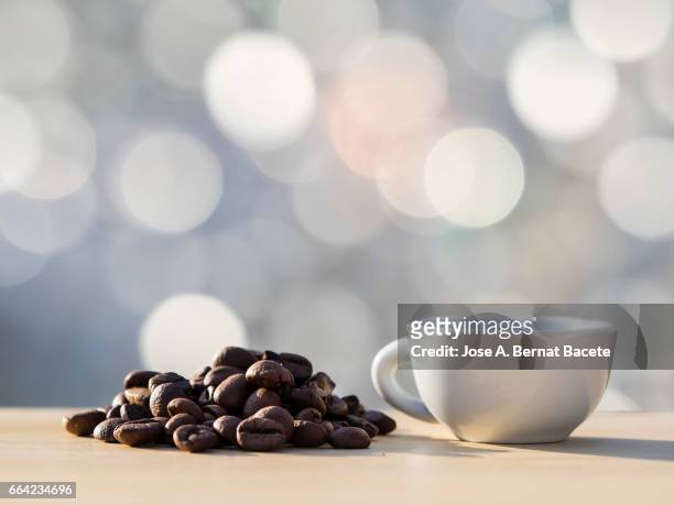 heap of coffee beans toasted and a cup of coffee  on a table of wood illuminated by the light of the sun - frescura stock pictures, royalty-free photos & images