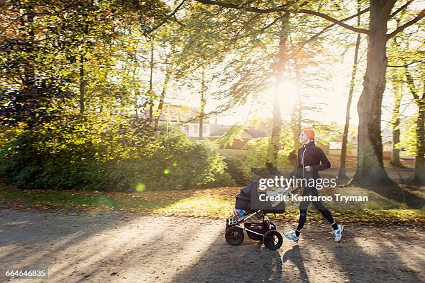 mother jogging with baby stroller on road against trees at park - jogging stroller stockfoto's en -beelden