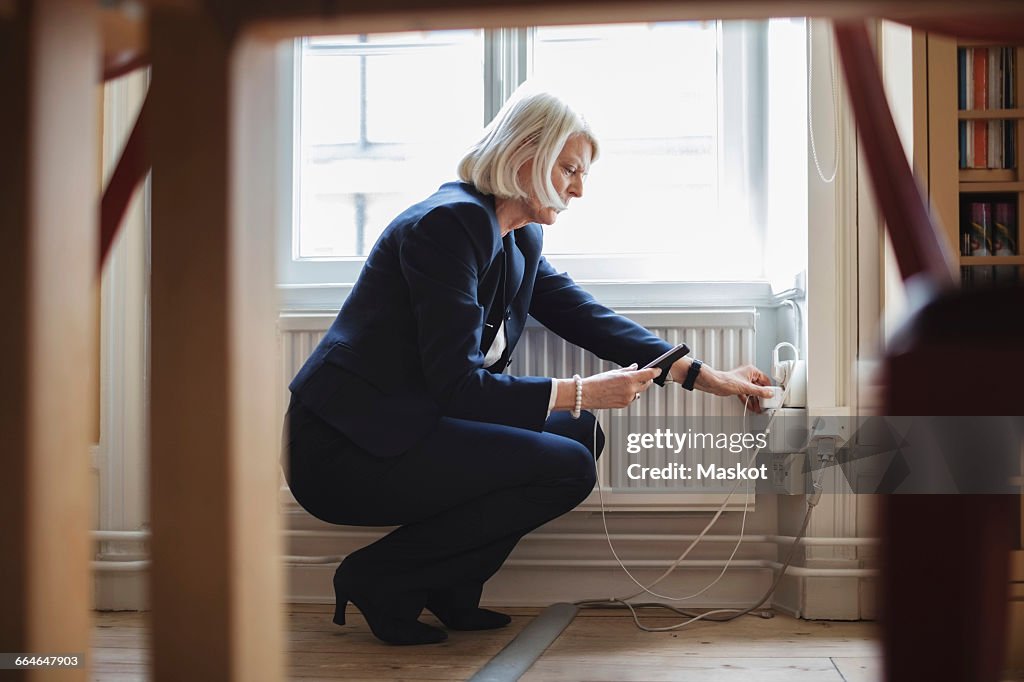 Senior woman crouching while charging mobile phone by radiator with furniture in foreground at library