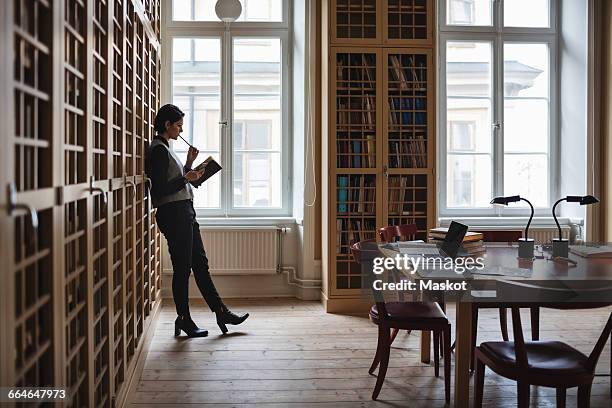 thoughtful lawyer holding book while leaning on shelf in library - studio legale foto e immagini stock