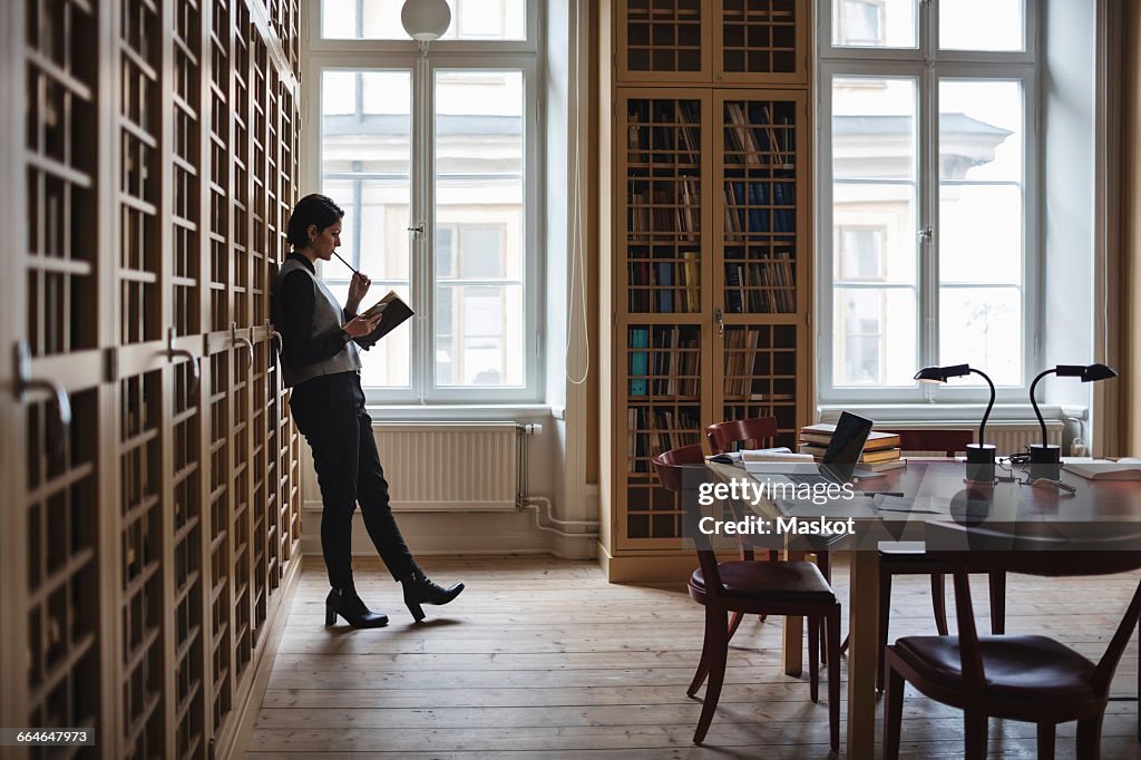 Thoughtful lawyer holding book while leaning on shelf in library