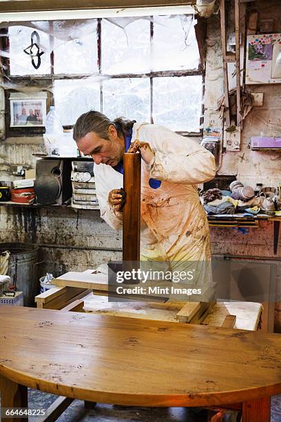 man standing in a carpentry workshop, wearing protective suit and gloves, applying varnish onto a wooden surface with a cloth. - wooden surface finishes foto e immagini stock