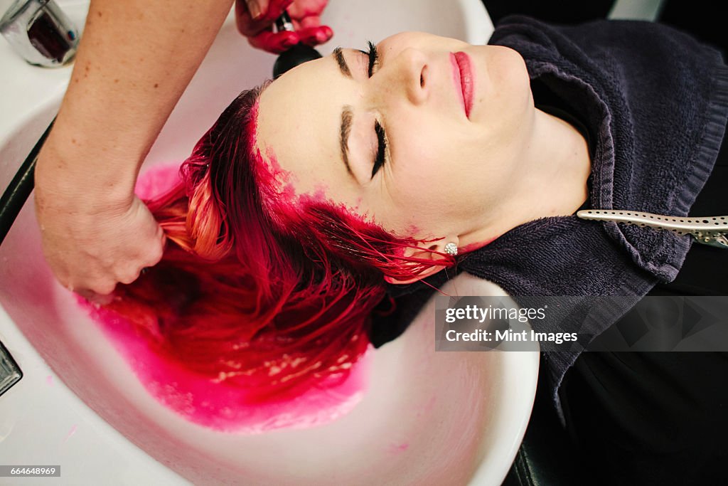 A hair salon client having red hair dye rinsed from her hair over a basin.
