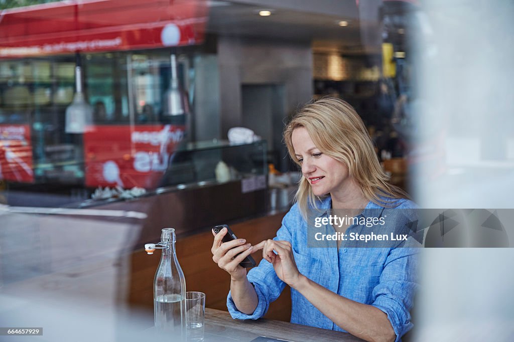 Mature woman sitting in cafe, using smartphone, bus reflected in window